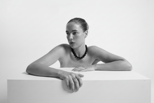 Load image into Gallery viewer, Woman with a beaded necklace sitting on a white surface against a plain background